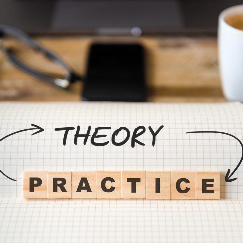 The photo shows a checkered block with the written word "theory". The word "practice" is also depicted with wooden stones placed on top. The two words are connected by arrows. In the background you can see an office desk blurred.